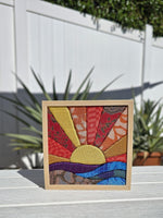 May include: A square fabric art piece framed in light wood, featuring a sunburst design with rays in yellow, orange, and red patterned textiles. Below the sun, horizontal strips of purple, blue, and teal fabric represent a landscape.