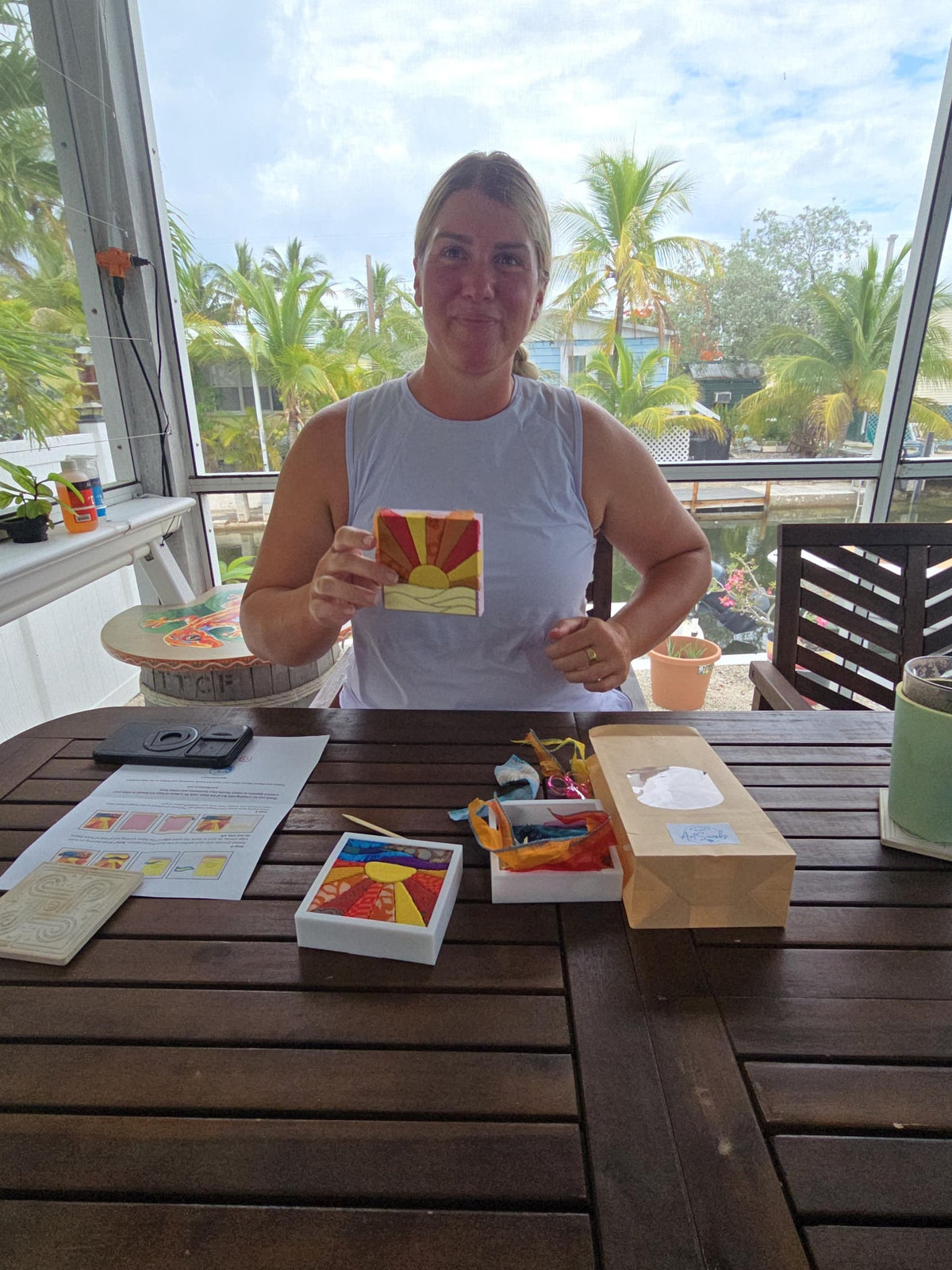 May include: A small rectangular art piece featuring a stylized sun rising over waves in shades of yellow, orange, and red. The design is composed of geometric segments with textured patterns. The piece is displayed on a table alongside a paper bag.