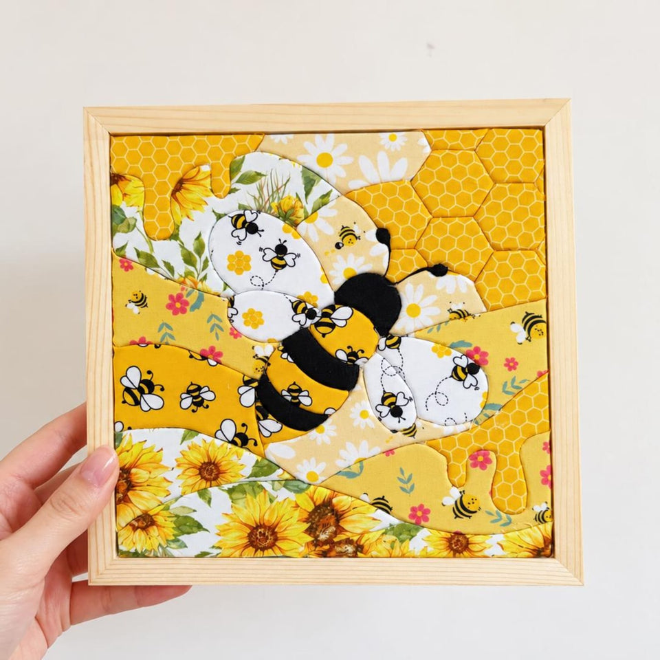 May include: A square fabric wall art piece in a light wood frame featuring a central bumblebee appliqué. The design incorporates yellow and white patterned fabric sections with sunflowers, daisies, honeycomb motifs, and small bee illustrations.