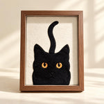May include: A framed wall art piece featuring a black cat silhouette with bright orange eyes and a long, upright tail. The cat is rendered in a textured black material against a cream-colored background, enclosed in a simple brown wooden frame.