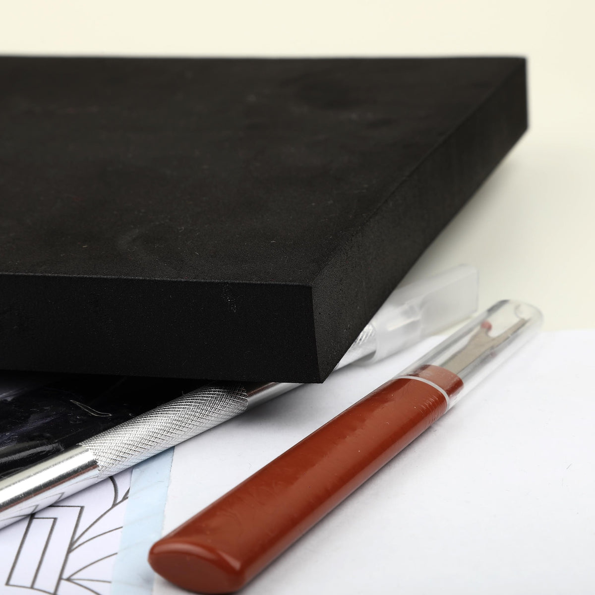 May include: A thick rectangular black foam block positioned above a silver metal craft knife with a textured handle and a brown seam ripper featuring a clear plastic protective cap, resting on a white surface with printed line art.