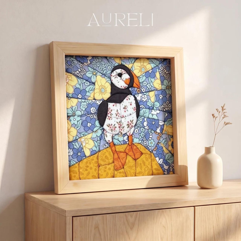 May include: A square framed textile art piece featuring a puffin with a floral patterned body, standing on a yellow textured base. The background consists of blue and yellow floral fabric segments. The word AURELI is printed at the top.