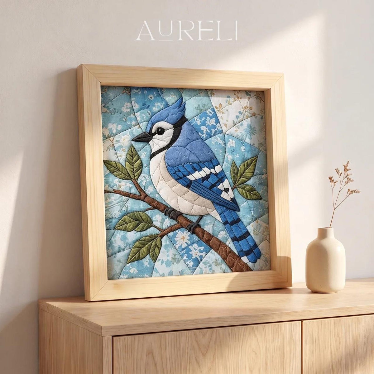 May include: A square framed textile art piece featuring a blue jay perched on a brown branch. The bird is crafted from blue, white, and black fabric patches. The background consists of light blue floral patterned fabric squares. The text AURELI is visible.
