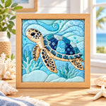 May include: A square framed fabric wall art piece featuring a quilted sea turtle swimming through blue waves. The turtle has a tan body with dark spots and a shell composed of various blue patterned fabric patches, surrounded by light blue coral.