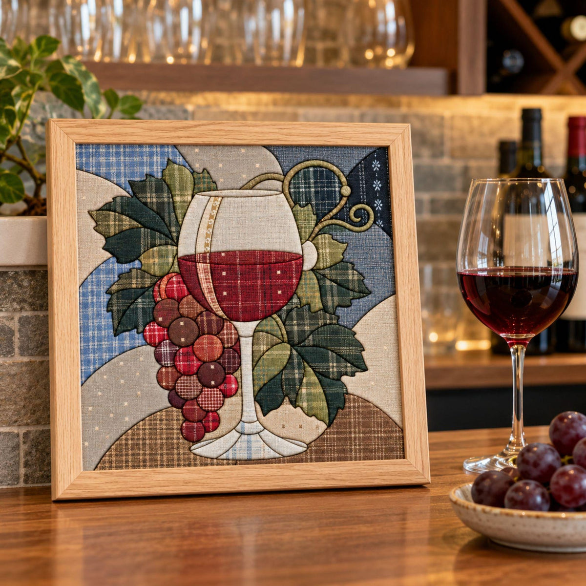 May include: A square framed fabric wall art piece featuring a quilted applique design of a wine glass filled with red liquid and a cluster of red grapes. The artwork uses various plaid and patterned fabric patches in shades of blue, green, and brown.