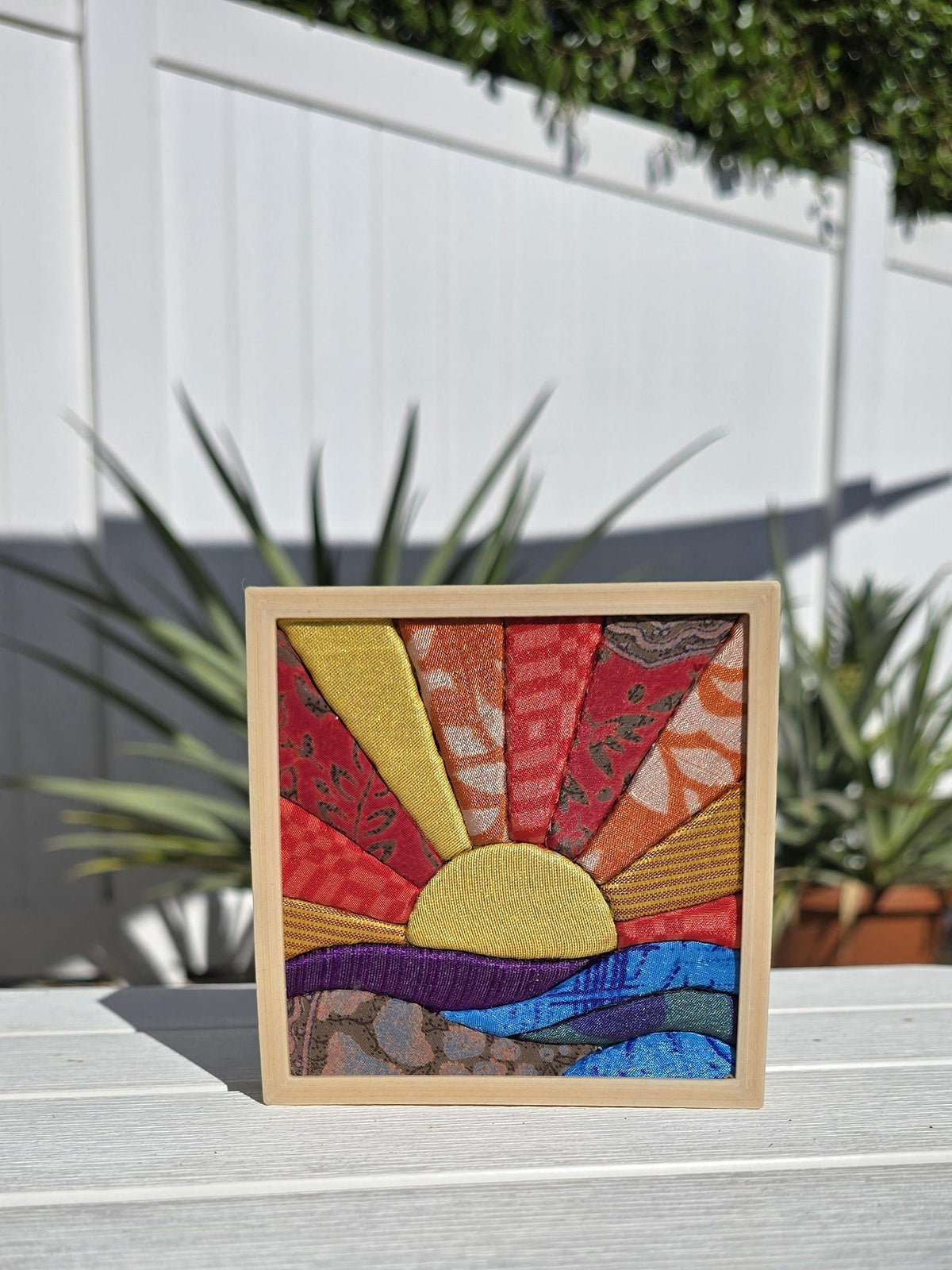 May include: A square fabric art piece framed in light wood, featuring a sunburst design with rays in yellow, orange, and red patterned textiles. Below the sun, horizontal strips of purple, blue, and teal fabric represent a landscape.
