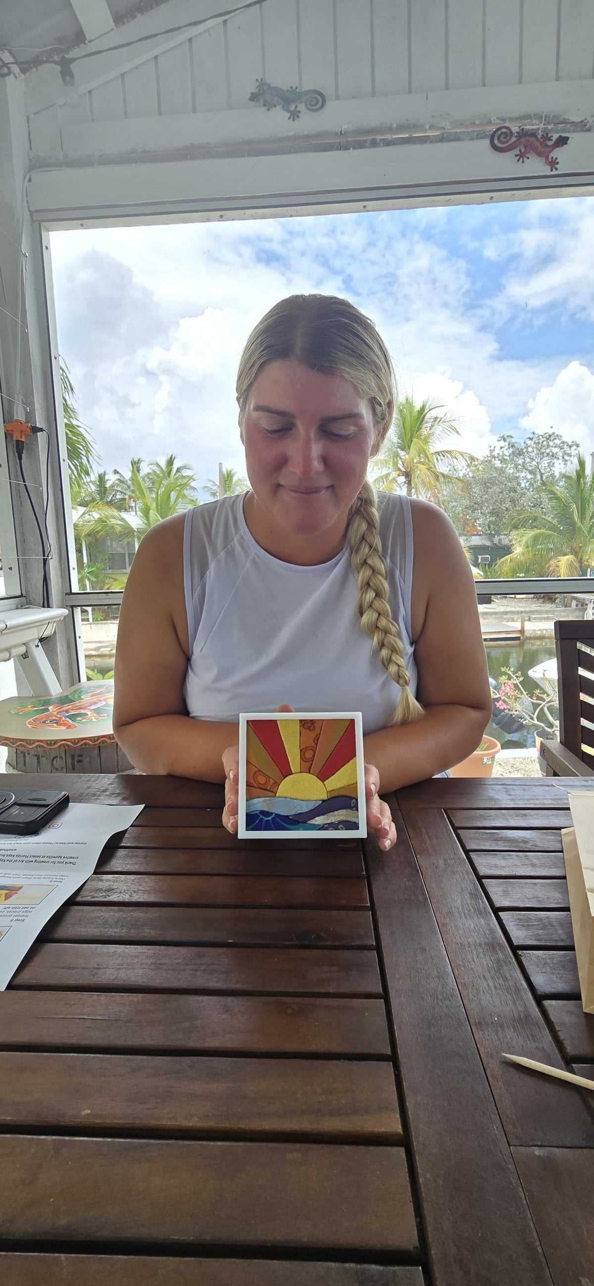 May include: A square decorative tile featuring a vibrant mosaic-style design of a yellow sun rising over blue ocean waves. The sun rays are depicted in alternating shades of red, orange, and yellow, framed by a clean white border.