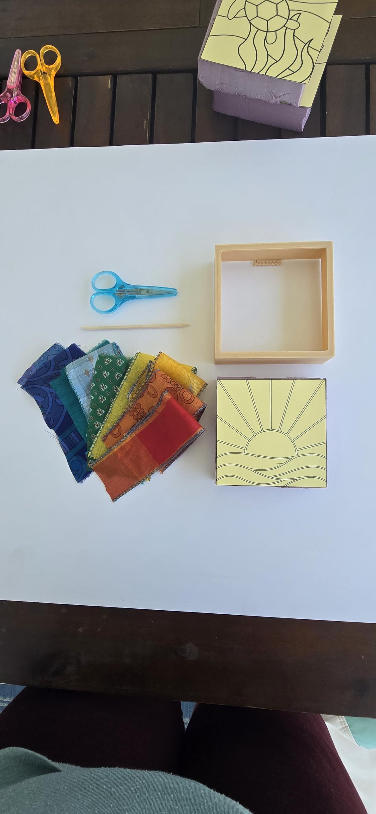 May include: A craft kit containing a square wooden frame, a yellow template with a sun and horizon line design, a wooden stylus, blue scissors, and a fan of fabric strips in shades of blue, green, yellow, orange, and red on a white surface.