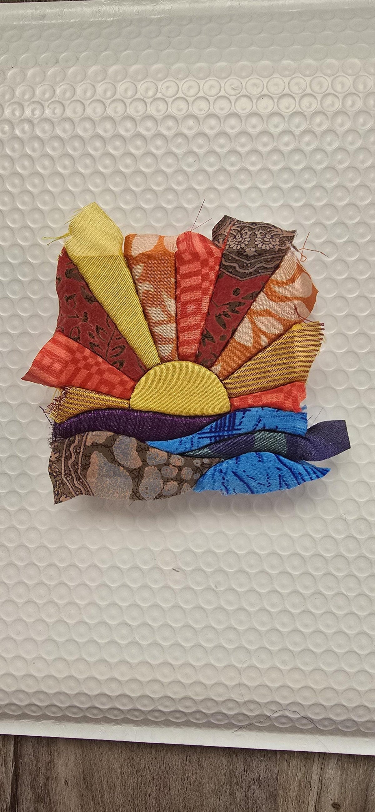 May include: A fabric applique depicting a sunrise over water, constructed from various textile scraps in shades of yellow, orange, red, brown, purple, and blue. The design features a central yellow sun semicircle with radiating fabric rays.