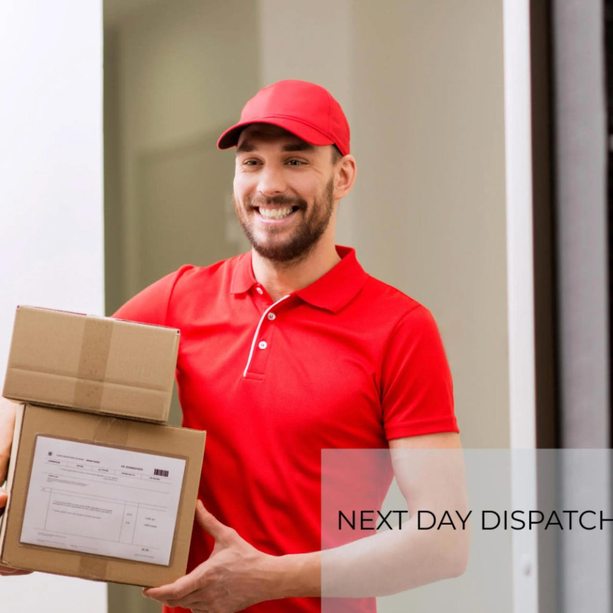 May include: A delivery worker wearing a bright red polo shirt and a matching red baseball cap, holding two stacked brown cardboard shipping boxes. The bottom box features a white shipping label with printed text and a barcode.