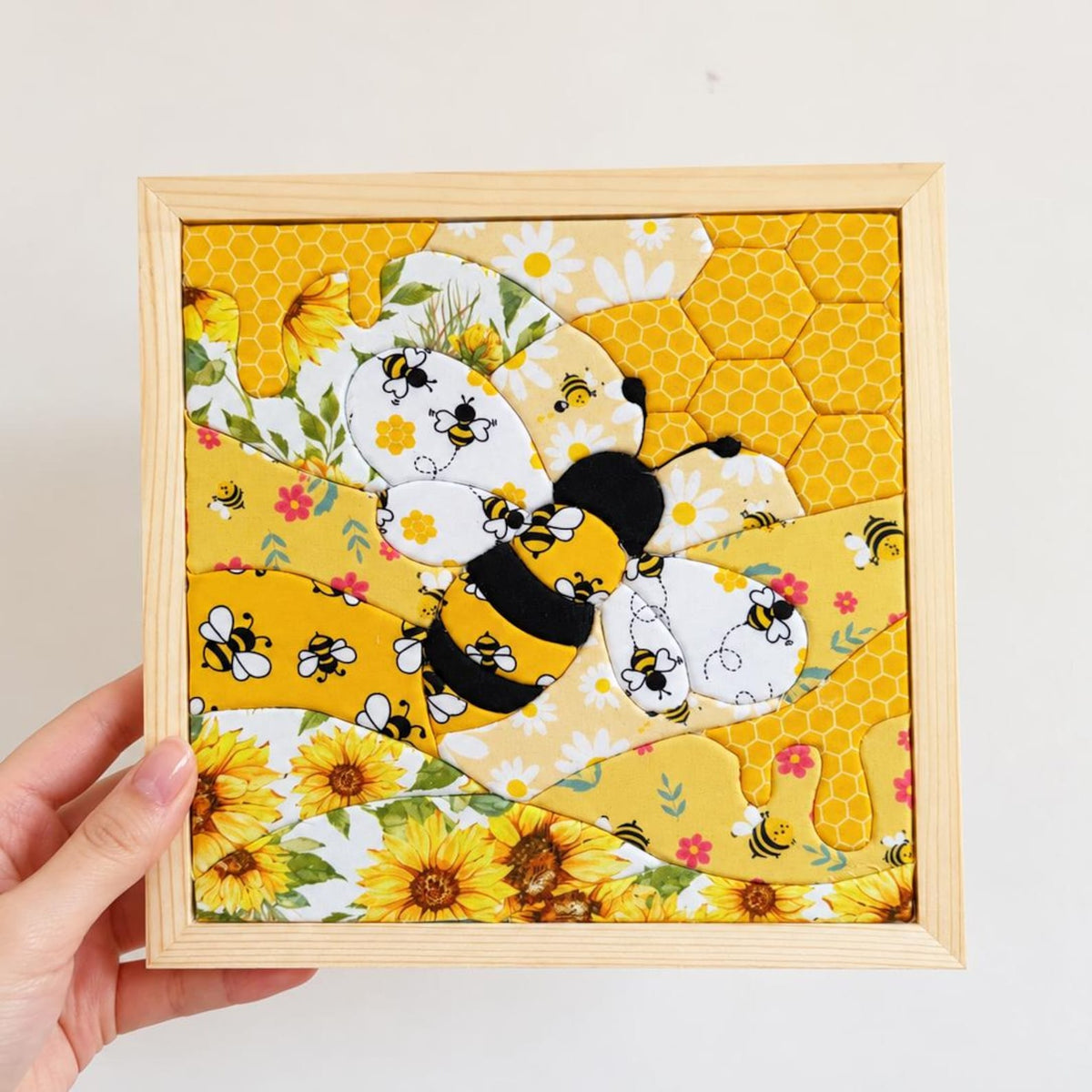 May include: A square fabric wall art piece in a light wood frame featuring a central bumblebee appliqué. The design incorporates yellow and white patterned fabric sections with sunflowers, daisies, honeycomb motifs, and small bee illustrations.