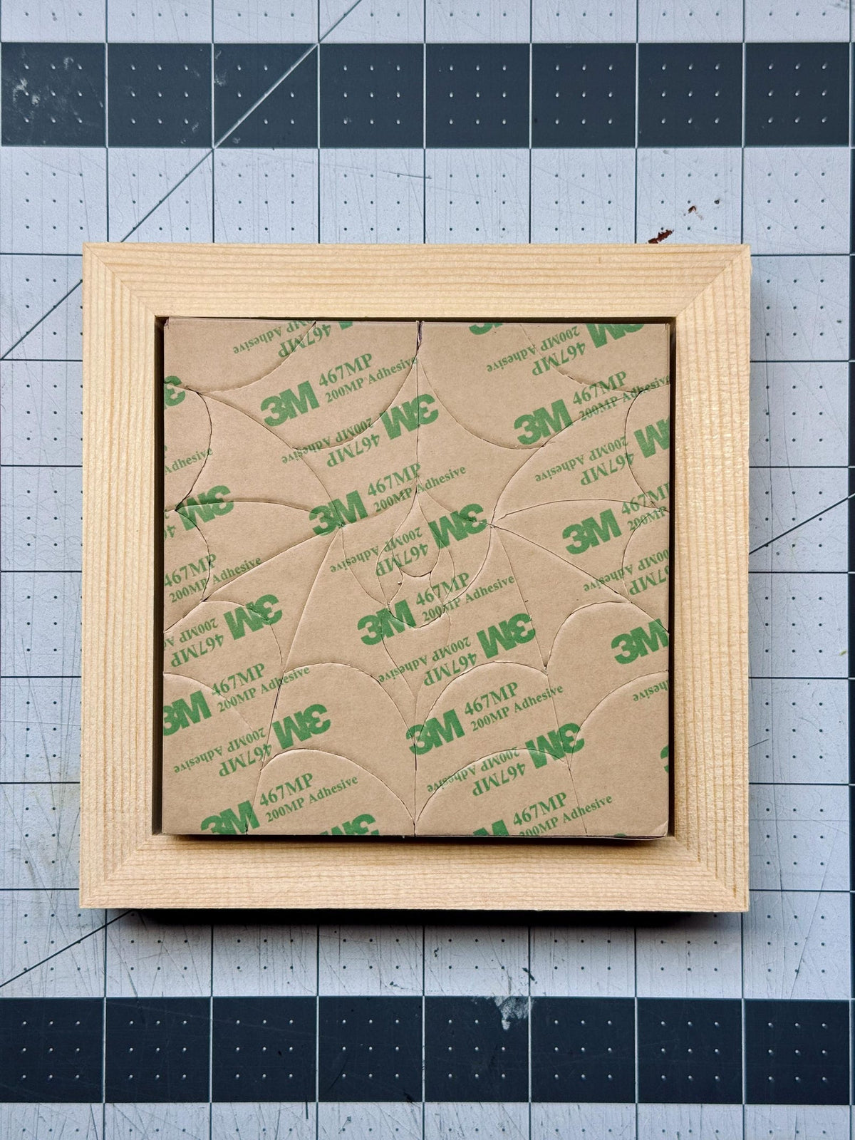 May include: A square light-colored wooden frame containing a series of interlocking tan adhesive-backed pieces. The surface of the pieces is covered in a repeating green 3M 467MP 200MP adhesive logo pattern, creating a geometric mosaic effect.