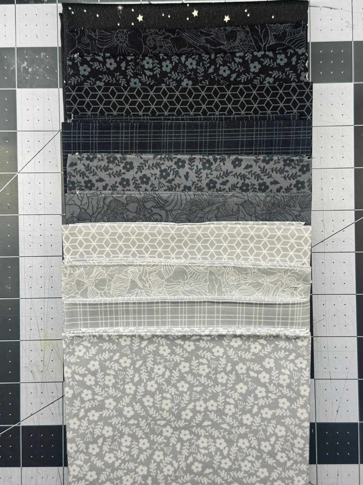 May include: A stack of ten square fabric swatches arranged in a gradient from black to light gray. The patterns include stars, geometric cubes, thin grid lines, and various floral designs printed in white or gray on solid black and gray backgrounds.