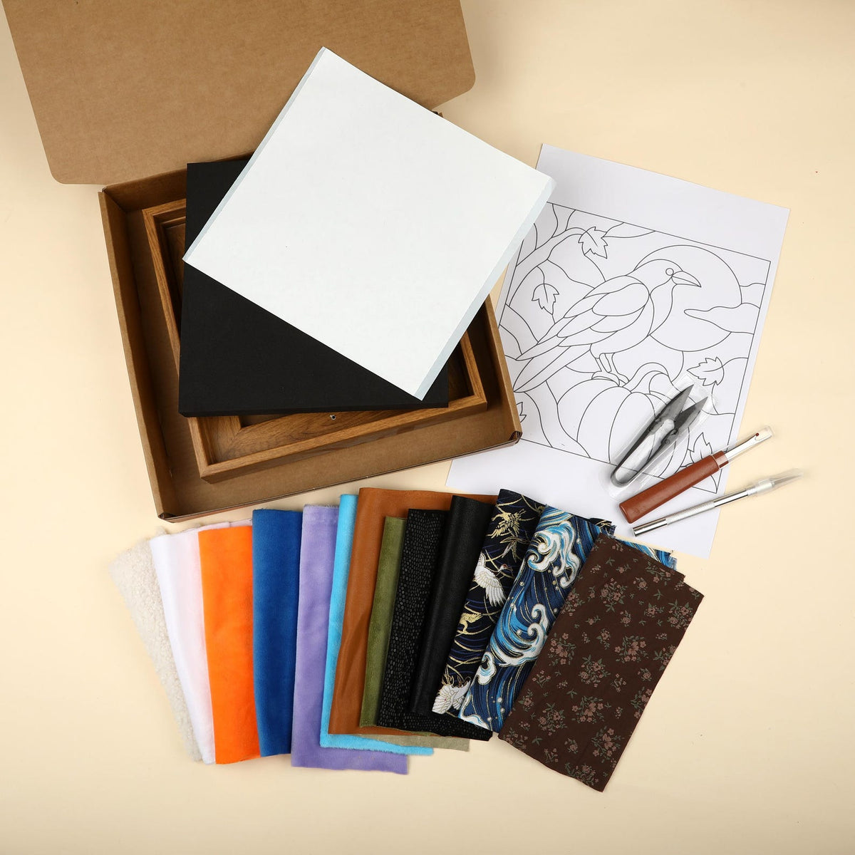 May include: A craft kit containing a brown cardboard box, a wooden frame, a white board, a black board, a paper template featuring a crow and pumpkin illustration, two metal craft tools, and a collection of fabric swatches in various colors and patterns.