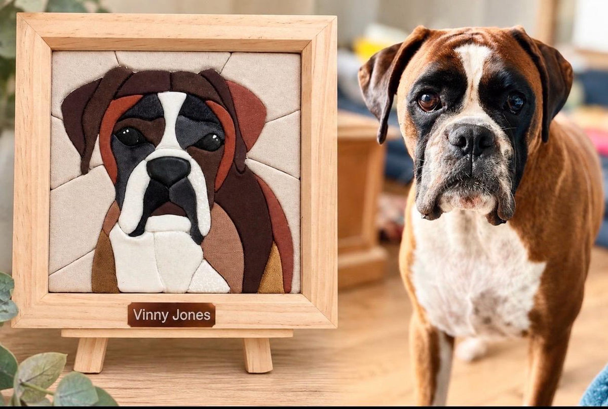 May include: A square wooden frame containing a mosaic-style portrait of a Boxer dog made from various colored fabric pieces. A small rectangular plaque at the bottom of the frame features the text Vinny Jones. A live Boxer dog stands nearby.