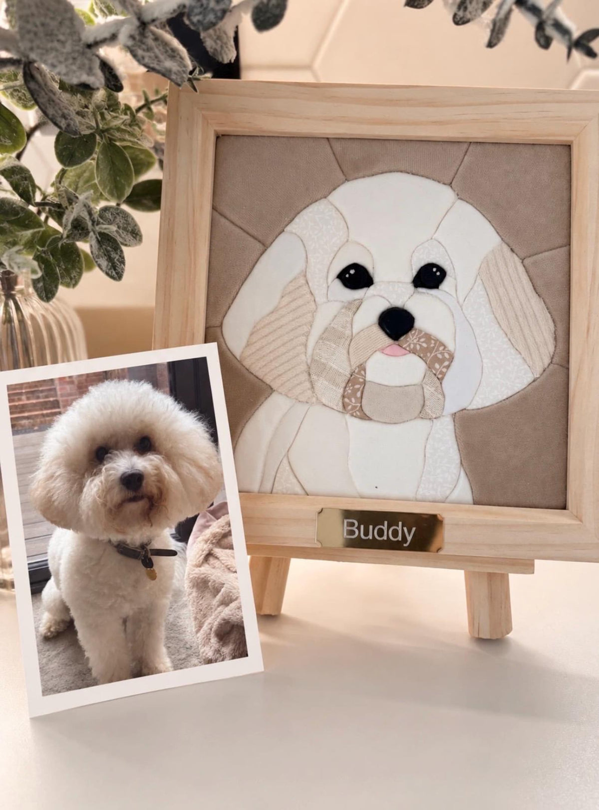 May include: A square fabric mosaic portrait of a white dog with floppy ears, framed in light wood. The artwork features various beige and white patterned fabric pieces and a gold-toned nameplate with the text Buddy. A photo of the dog is included.