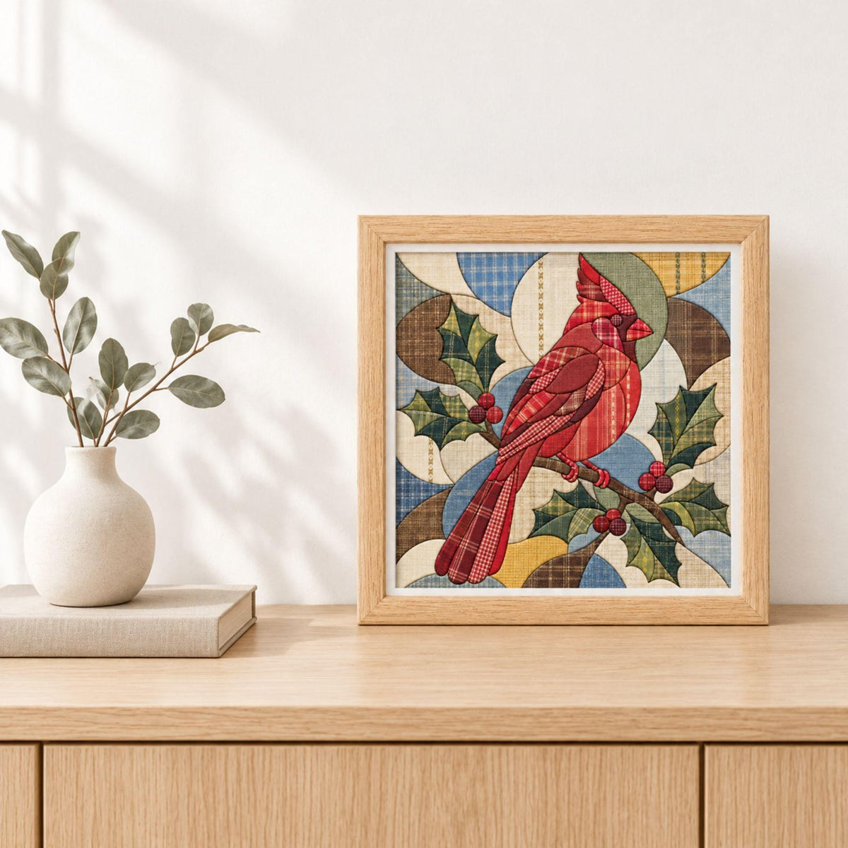 May include: A square framed wall art piece featuring a stylized red cardinal bird perched on a branch with green holly leaves and red berries. The design uses a patchwork quilt aesthetic with various plaid and solid fabric textures in red, blue, brown, and yellow tones.