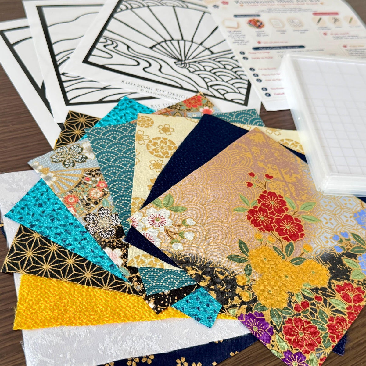 May include: A Kimekomi art kit featuring a collection of square fabric swatches with diverse patterns including floral, geometric, and wave designs in gold, blue, yellow, and white. Includes printed design templates and a white grid base.