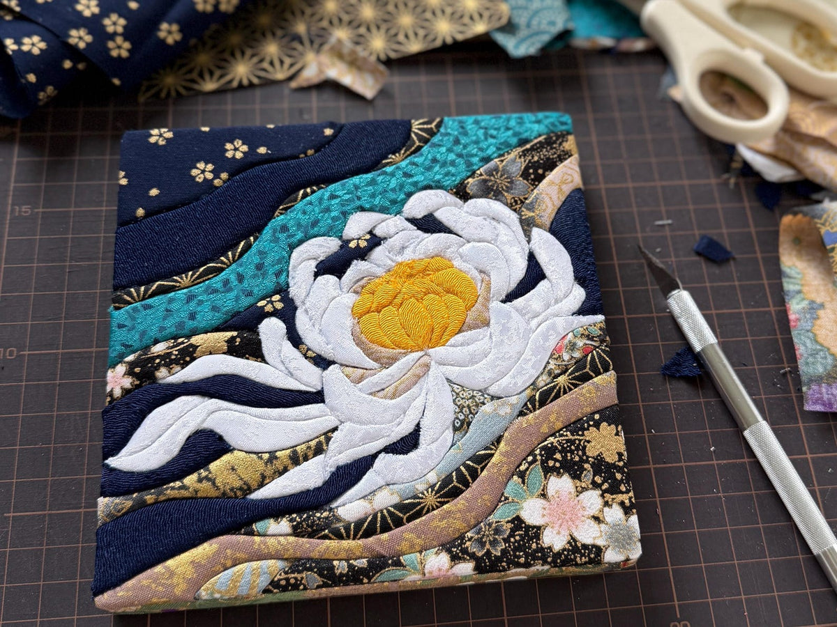 May include: A square fabric art piece featuring a central white lotus flower with a yellow center, surrounded by layered, wavy fabric strips in navy blue, teal, and gold-patterned prints with floral motifs on a dark background.