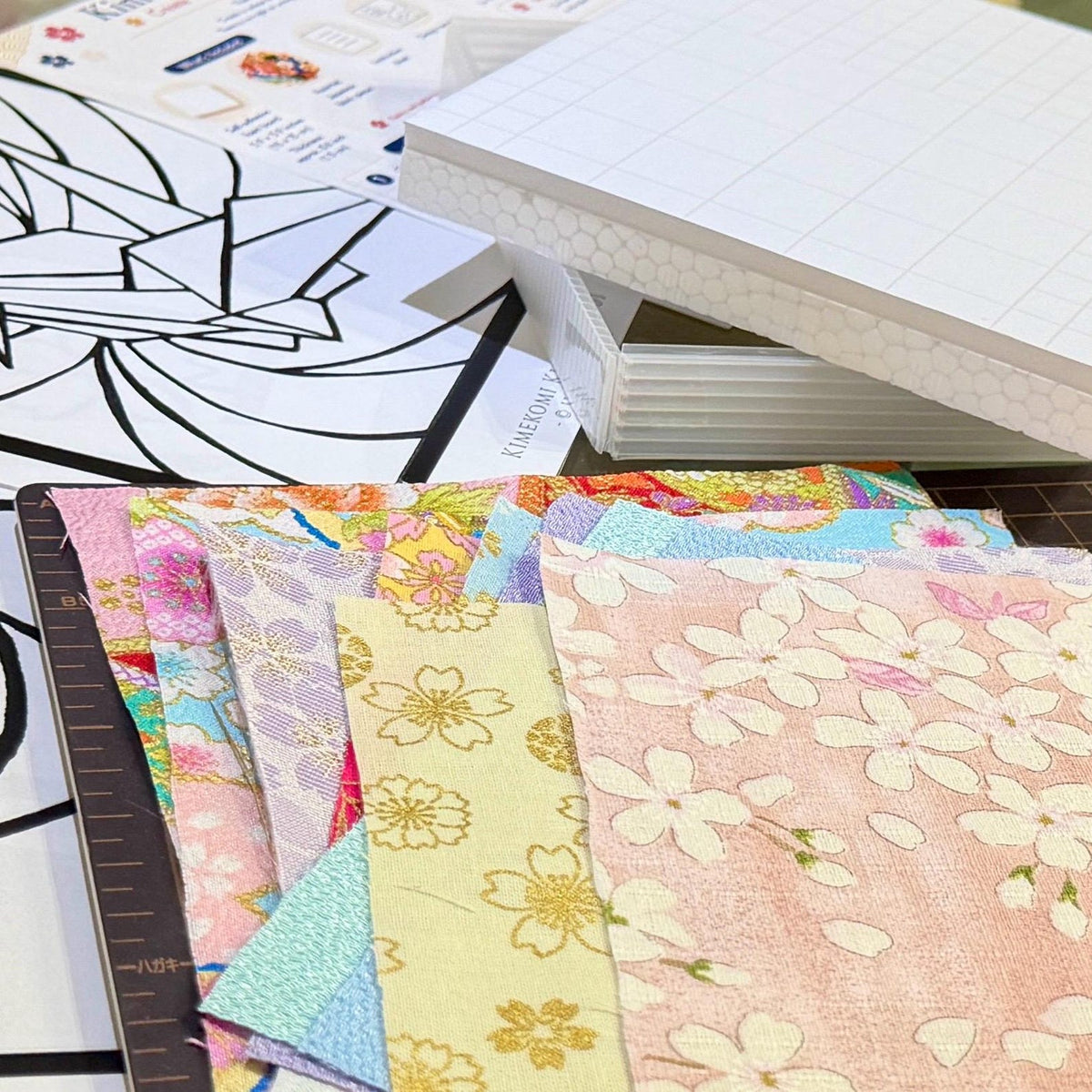 May include: A collection of rectangular fabric swatches featuring various pastel colors and floral patterns, including cherry blossoms. The fabric pieces are arranged in a stack next to a white grid-patterned foam board and a printed instruction sheet.