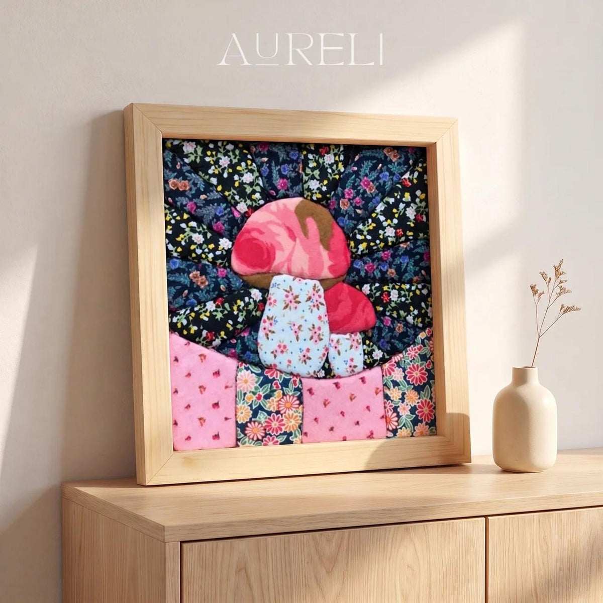 May include: A square wooden frame containing a fabric collage with the text AURELI at the top. The artwork features two mushrooms crafted from floral patterned fabric in shades of pink, blue, and black, set against a dark floral background.