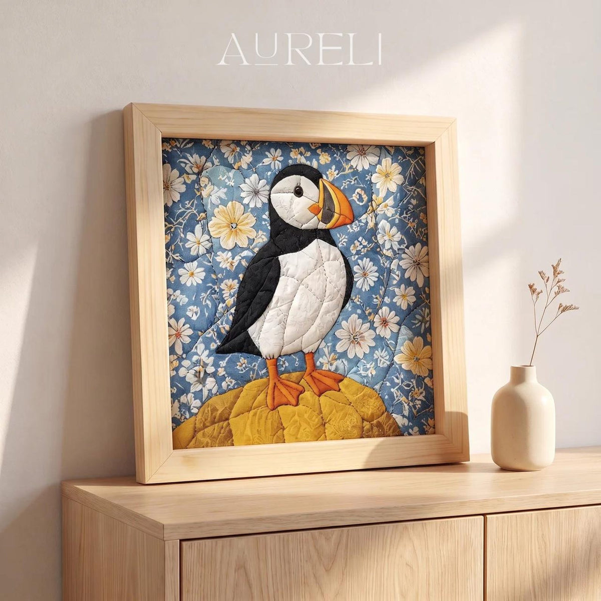 May include: A square framed textile art piece featuring a quilted puffin standing on a yellow rock against a blue floral background. The puffin has black and white feathers with an orange beak and feet. The word AURELI is printed above the frame.