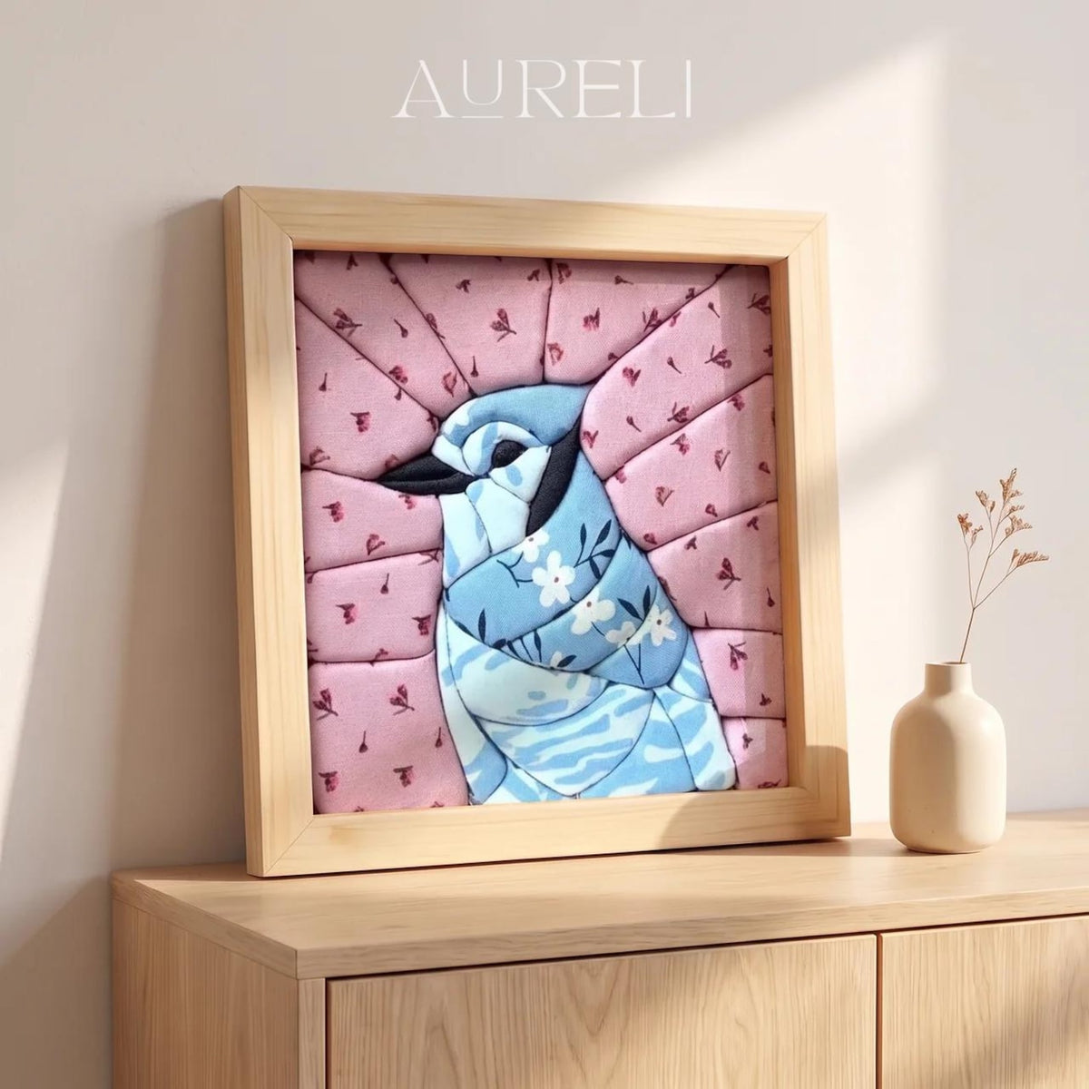May include: A square framed wall art piece featuring a blue jay bird illustration composed of segmented fabric panels. The bird is blue with white floral patterns, set against a pink background with small dark floral motifs. The text AURELI is visible.