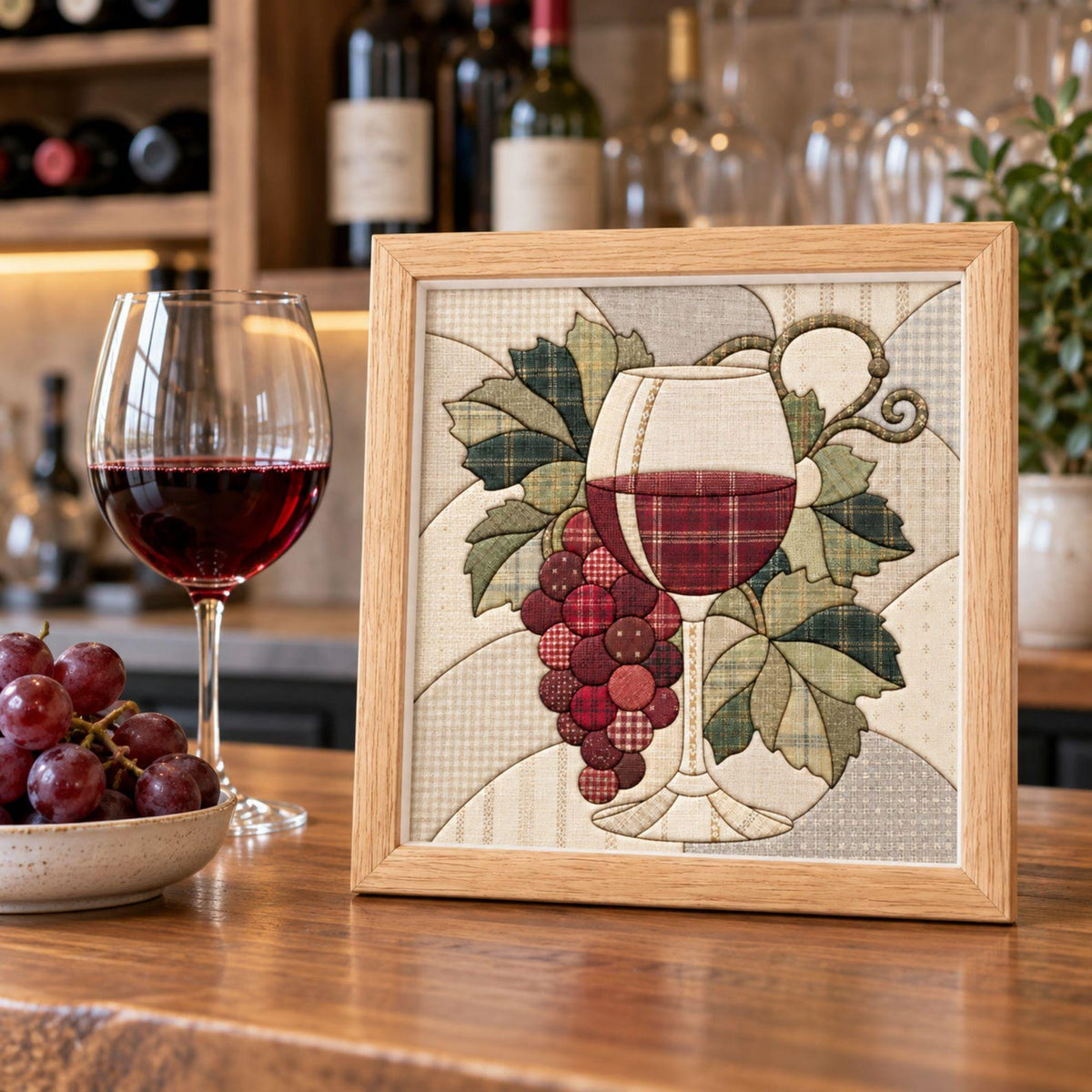 May include: A square framed fabric art piece featuring a quilted design of a wine glass filled with red liquid and a cluster of grapes with green leaves. The artwork uses various plaid and solid fabric textures in shades of red, green, and cream.